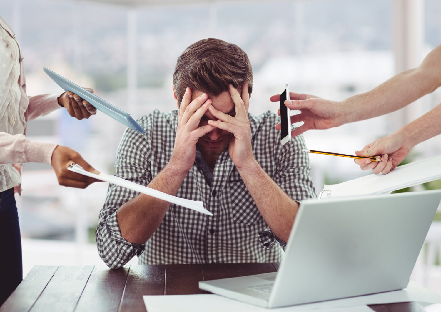 Homem sentado à mesa de trabalho com as mãos na cabeça, cercado por colegas oferecendo documentos e telefone, representando a necessidade de gerenciamento do estresse no ambiente profissional.