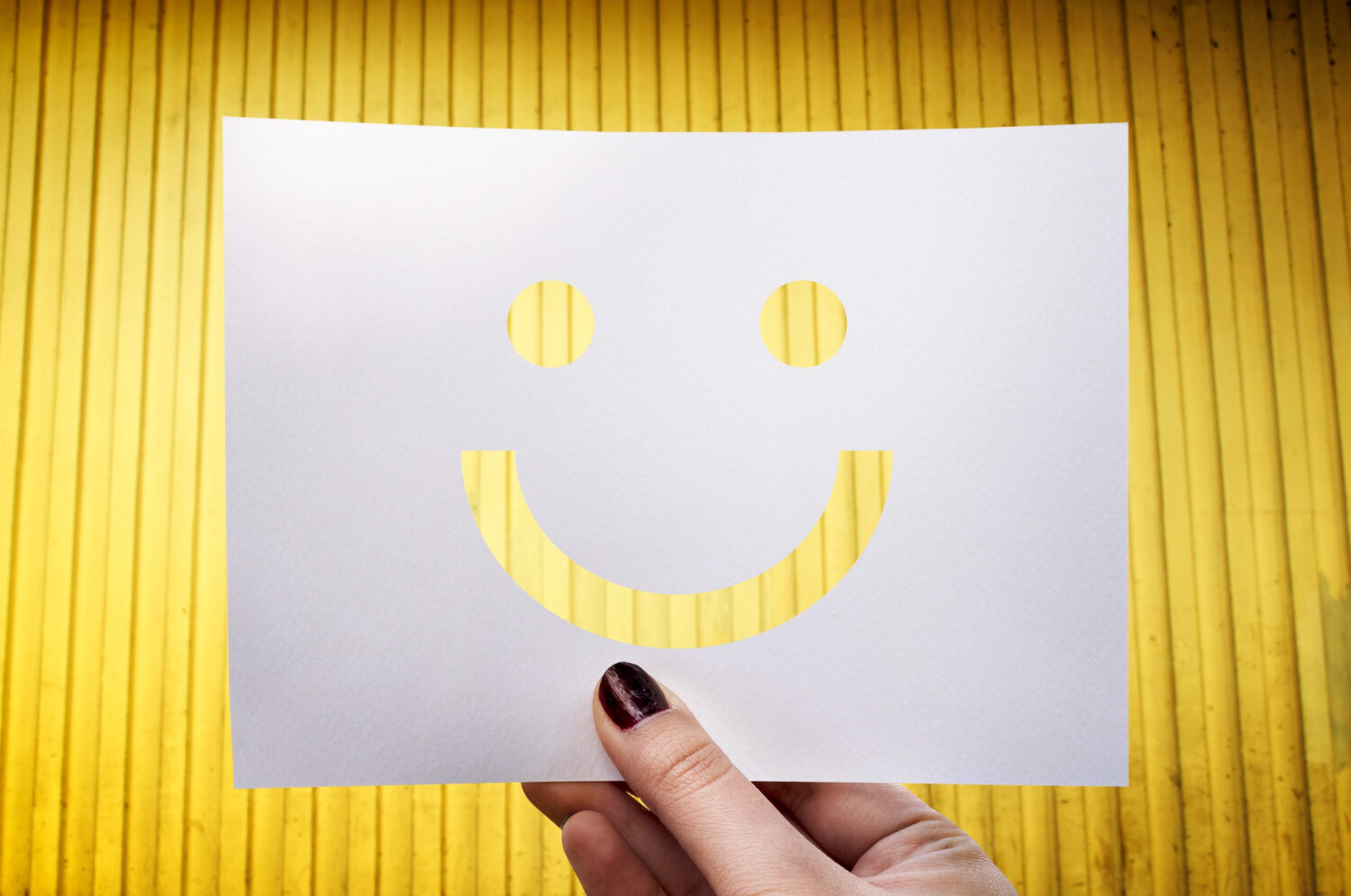 Mão segurando um papel branco recortado em formato de rosto sorridente, com fundo amarelo vibrante, simbolizando positividade e otimismo.